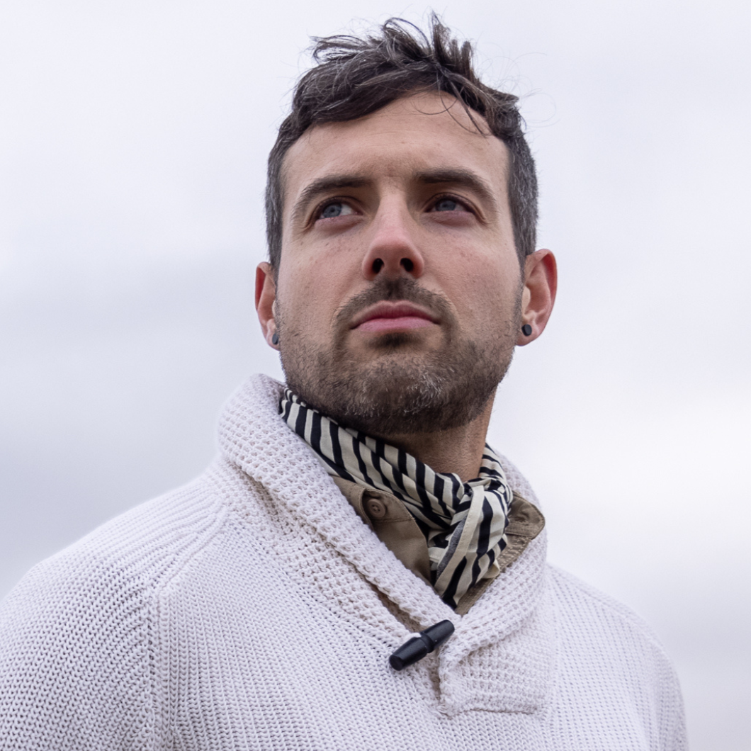 Man in a white sweater looking into the distance against a cloudy sky.