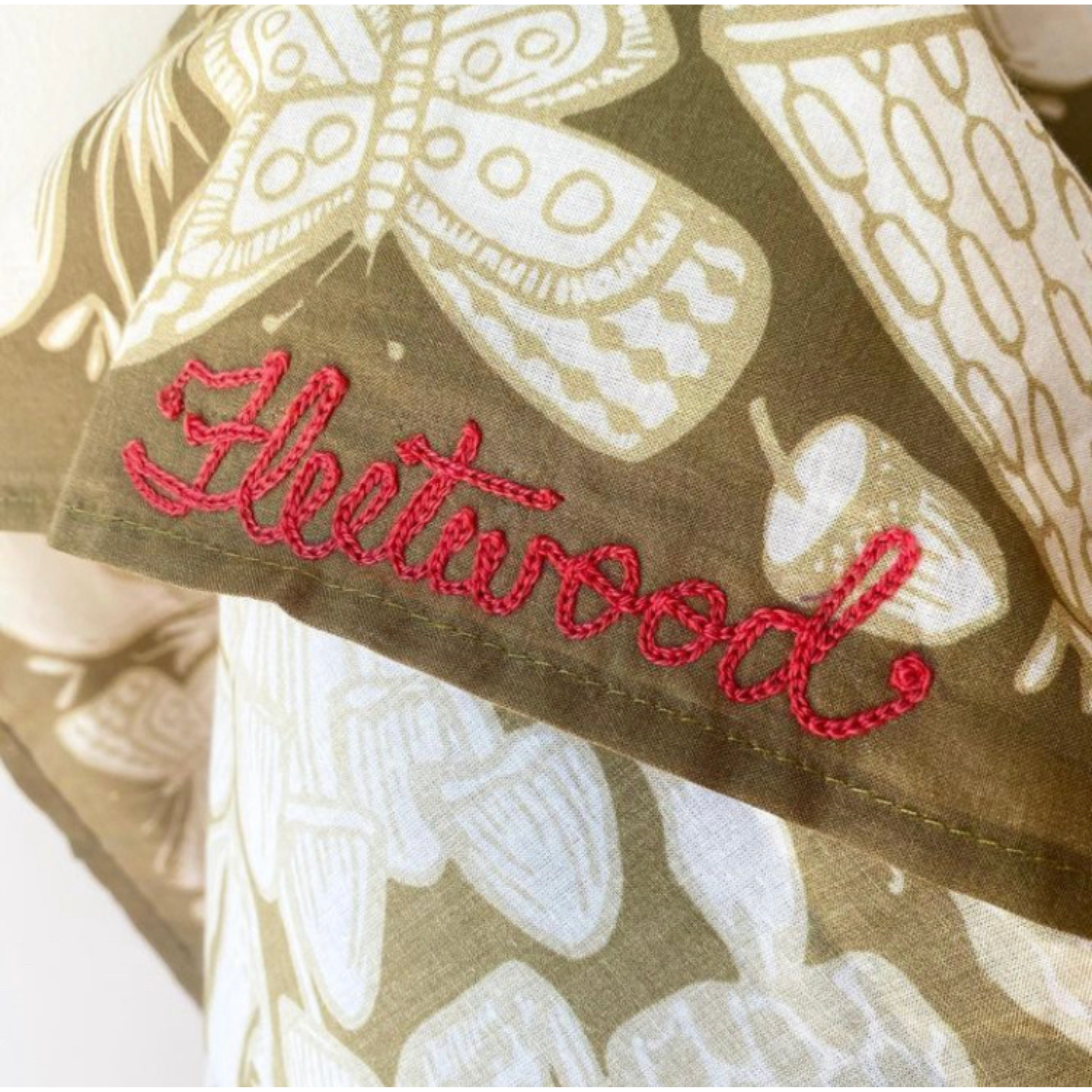 Close-up of folded olive-green bandana showing cream butterfly print and red embroidered Fleetwood on hem