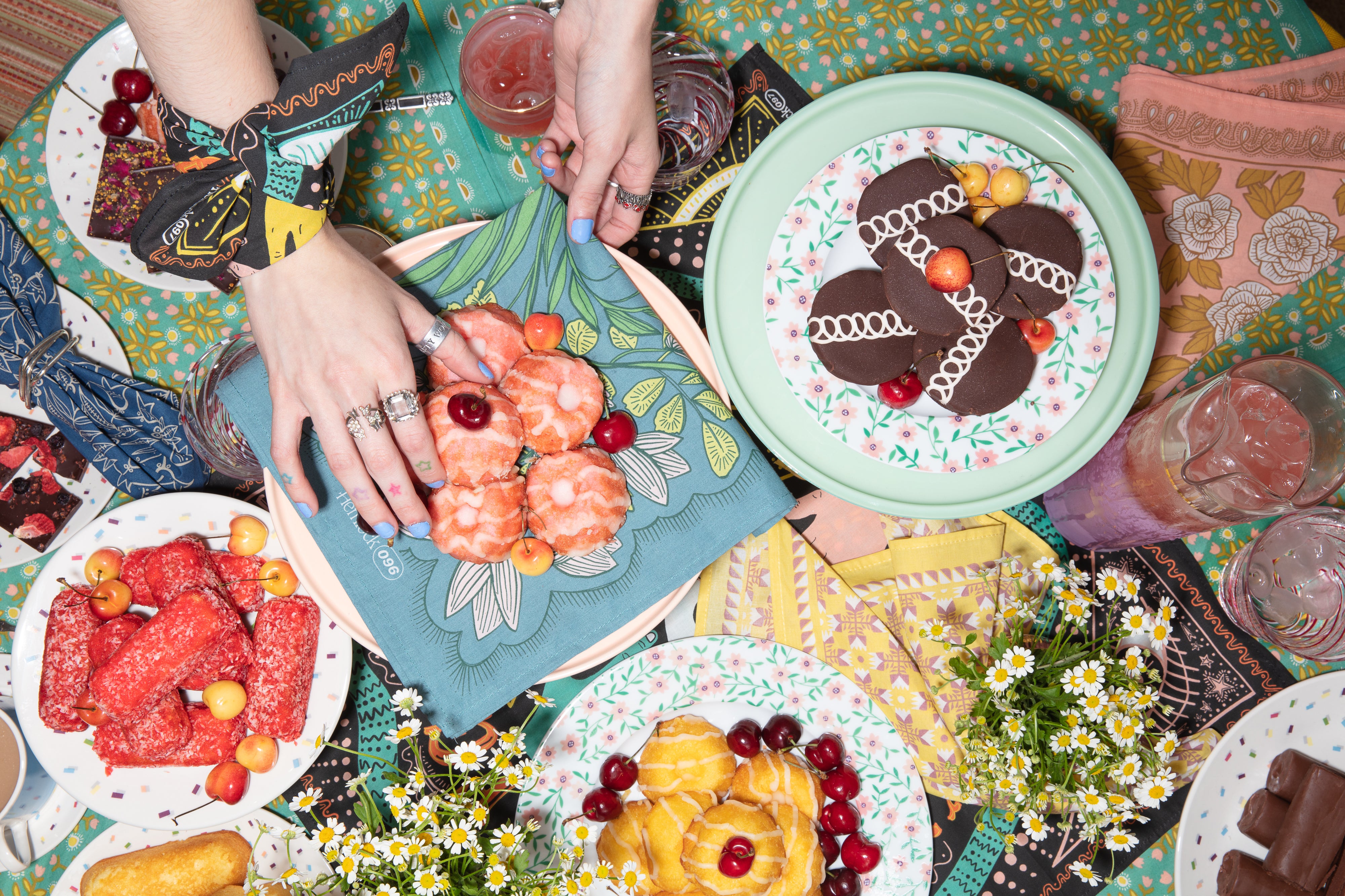 Hands arranging a plate of pastries on a colorful tablescape styled with Hemlock bandana napkins and mixed patterned tablecloth