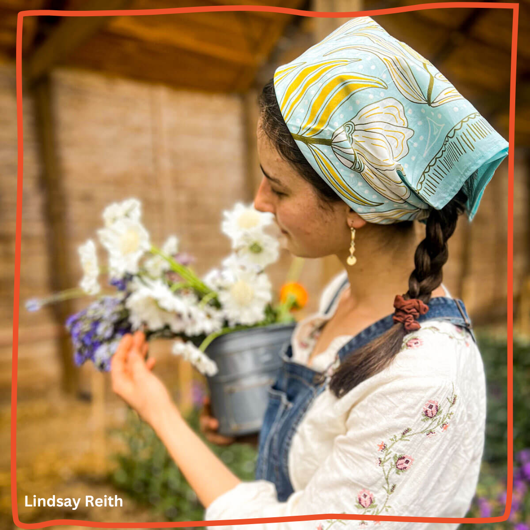 Young woman wearing a light blue floral bandana and denim overalls smelling white and purple flowers in a rustic barn