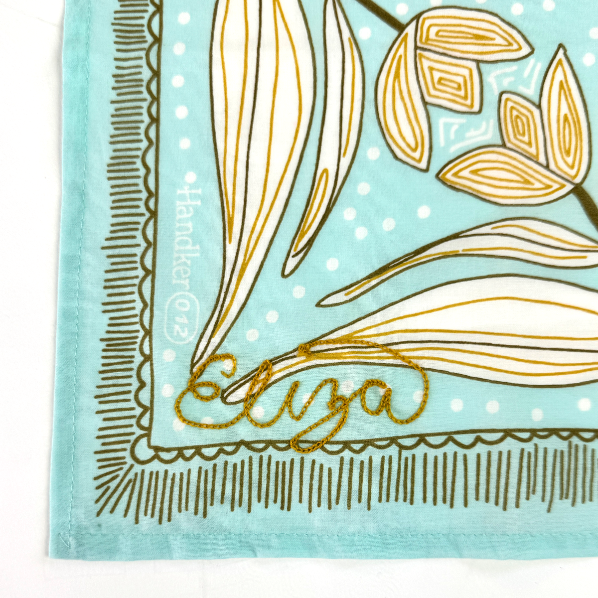 Turquoise bandana corner with white polka dots, botanical print and gold embroidered 'Eliza'; printed 'Handker©12' along left edge