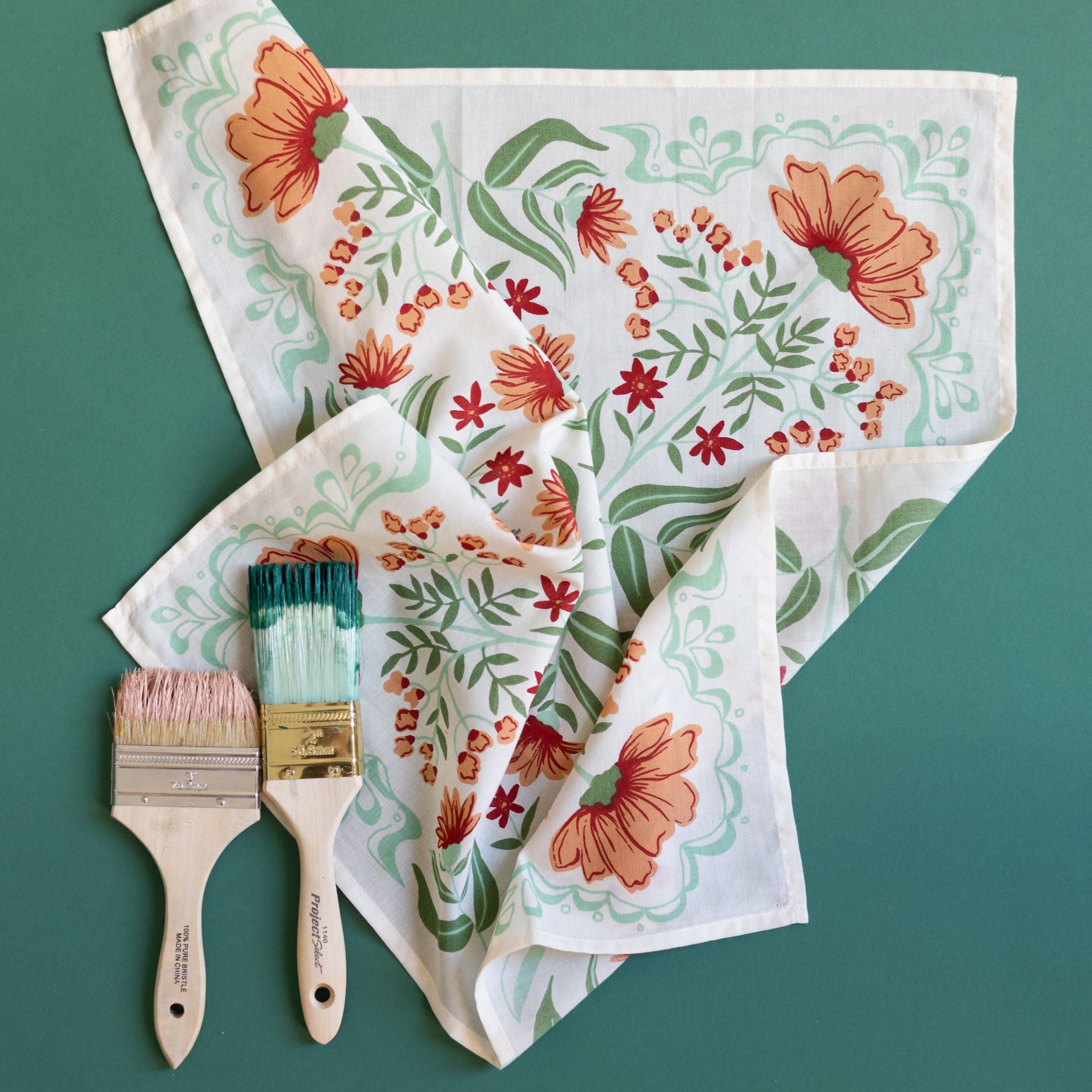 Flat lay of Flora bandana on green background showing white cotton with peach, burgundy and green floral print and two paintbrushes