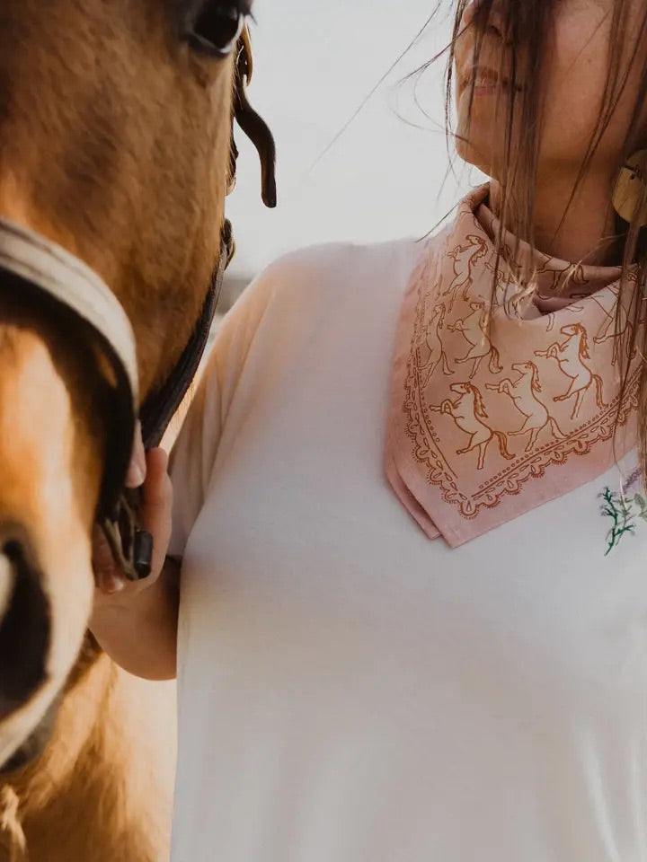 Woman wearing a pink bandana printed with galloping horses standing beside a brown horse in a close-up outdoor shot