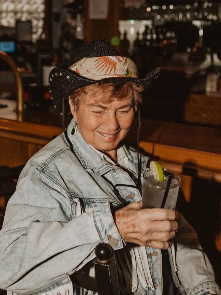 Older woman wearing tan floral bandana folded and tucked under a black straw hat, smiling in a dim bar while holding a drink