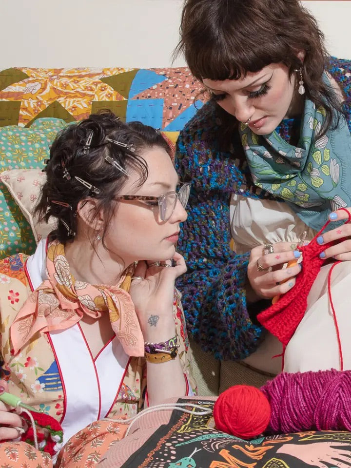 Two women knitting on a couch, one wearing a blue-pink Cassie bandana tied at her neck