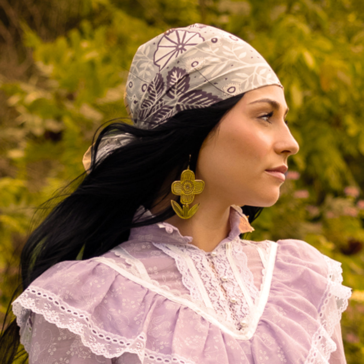 Woman in a lacy purple blouse with a patterned headscarf and gold earrings, standing outdoors with greenery in the background.