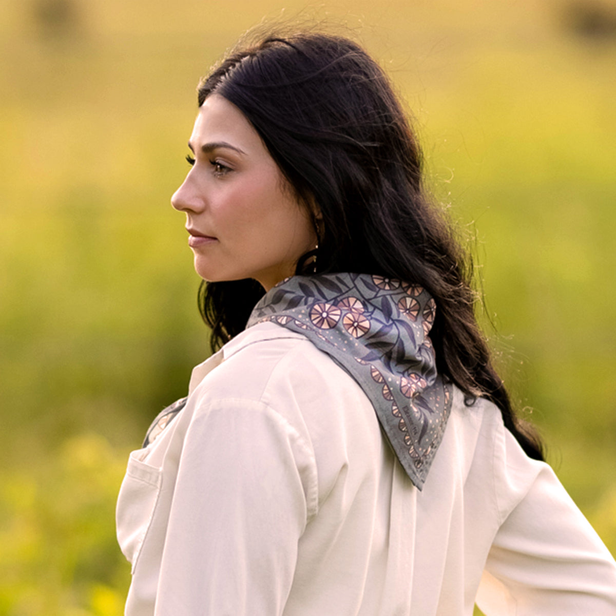 Woman in three-quarter profile wearing No. 114 Eleanor bandana tied at the neck, warm gray floral pattern in a blurred field