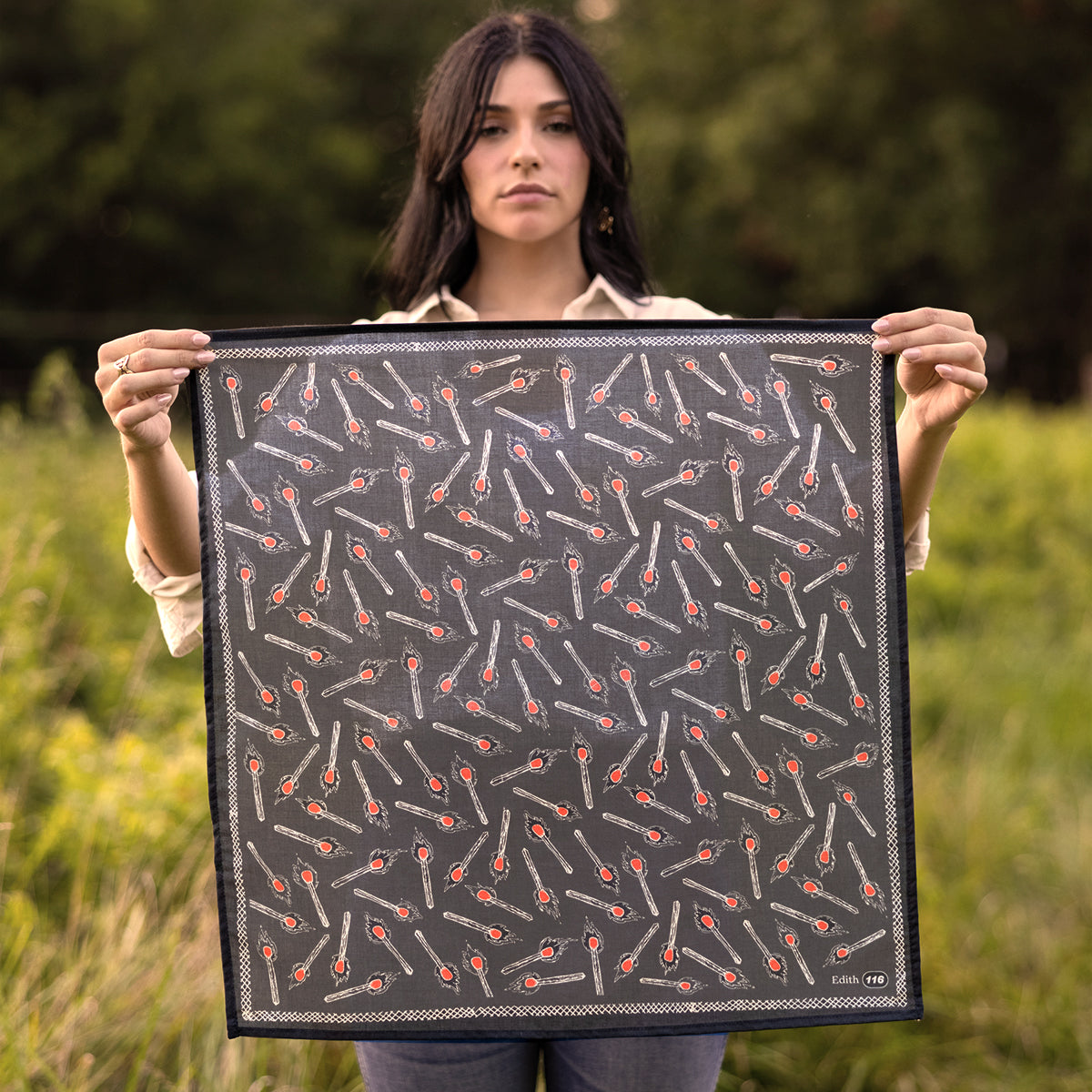 Front view of No. 116 Edith bandana showing charcoal gray background with spoon motif and orange and cream accents held by a person against a grassy outdoor background