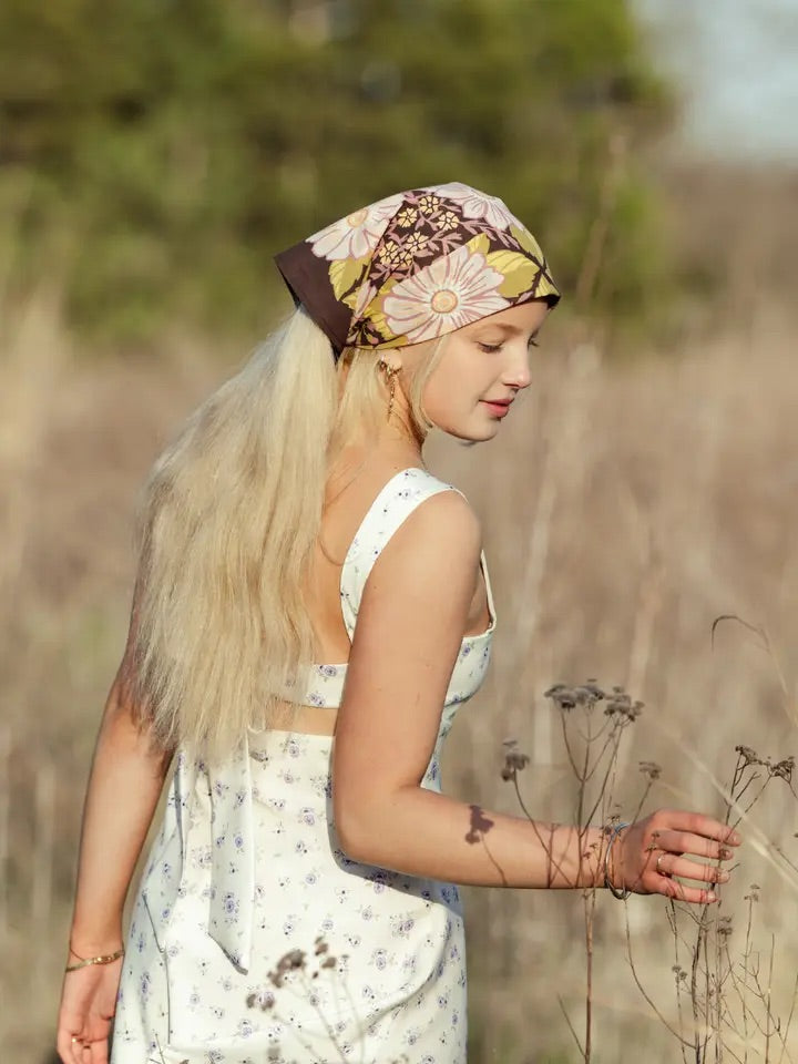 Side view of a blonde woman wearing a violet floral bandana tied at the back in a white sundress standing in a sunlit meadow