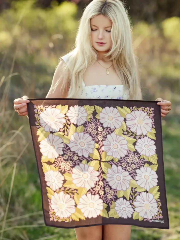 Woman holding a violet floral bandana flat, white‑pink daisy print with olive green leaves in a grassy outdoor setting