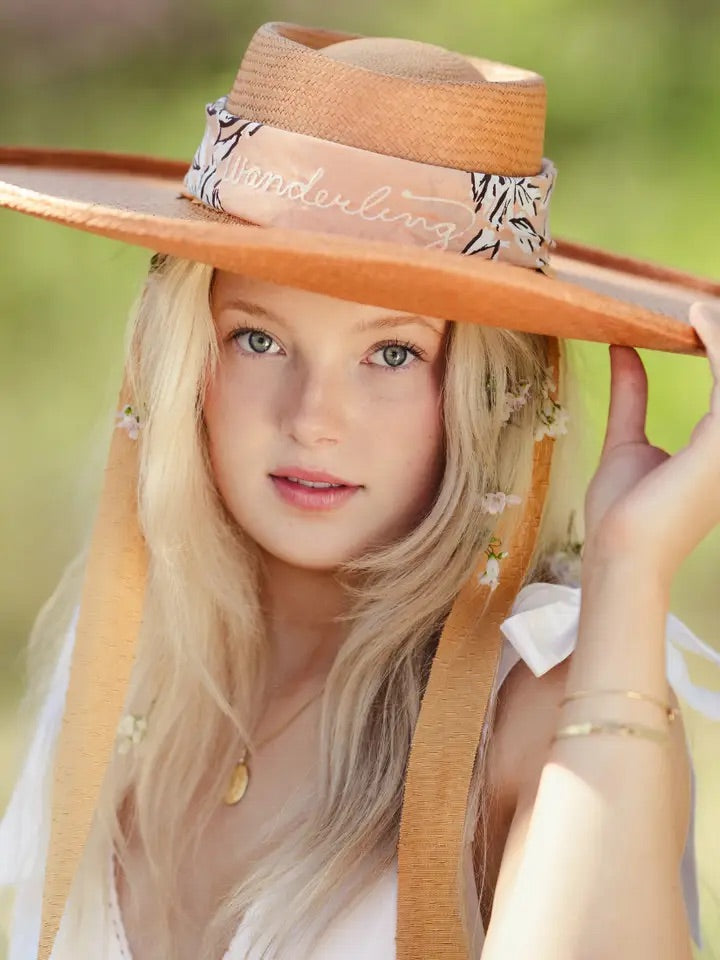 Young woman wearing a soft-peach floral bandana tied as a straw hat band showing the script "wandering", close-up outdoors