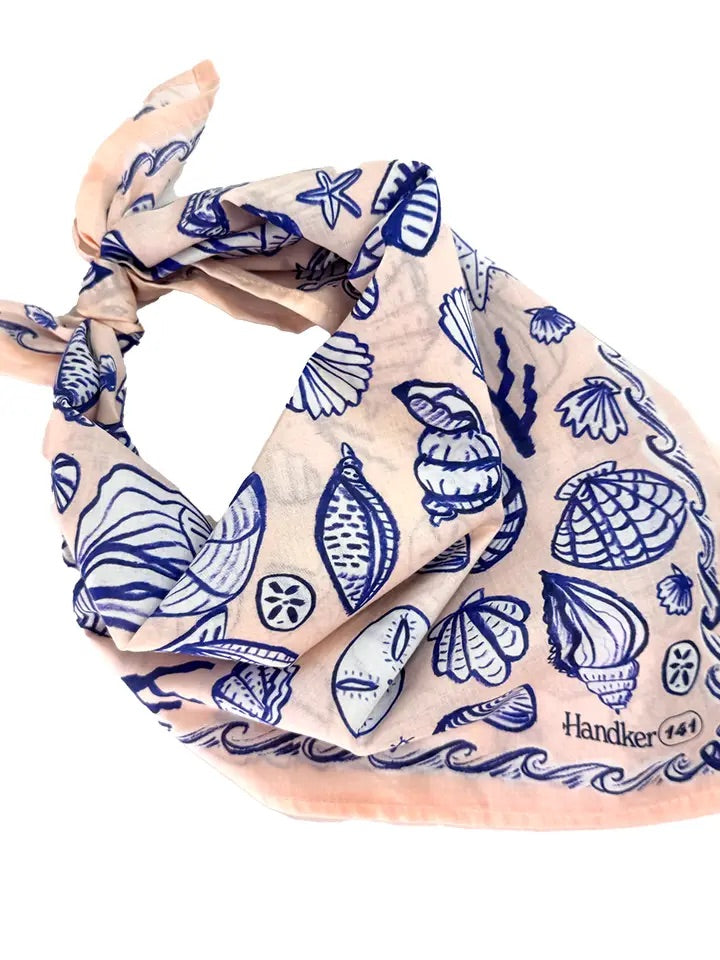 Angled view of No. 141 Seashore bandana showcasing pale peachy-pink cotton with bright blue seashell and starfish print, knotted on white background