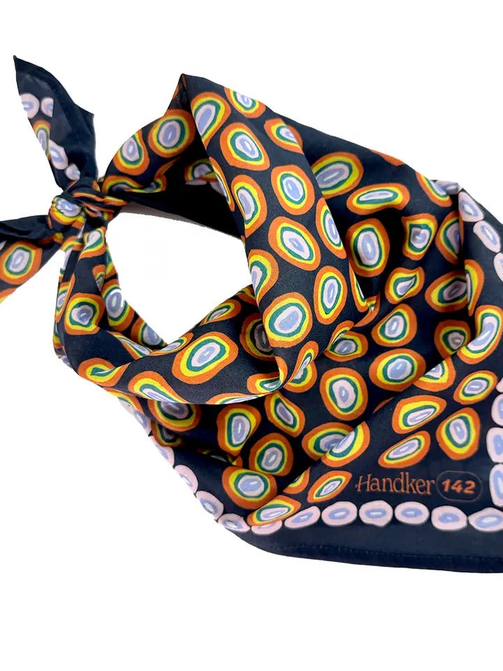 Angled view of No. 142 Lorena bandana showing orange, gold, and pink concentric ovals on navy blue, tied corner on white