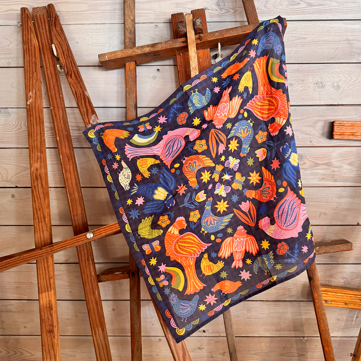 Colorful patterned bandana draped over a wooden easel against a wooden wall.