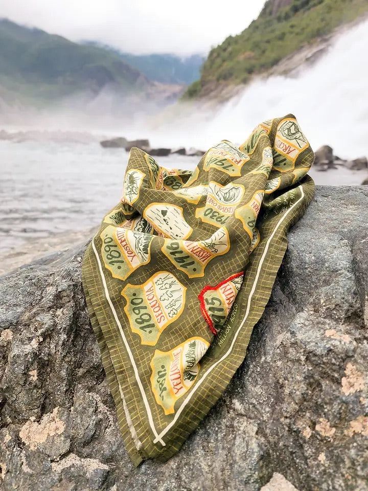 Draped green and beige National Parks bandana showcasing printed park badge illustrations on a rock with a misty river and mountains in the background