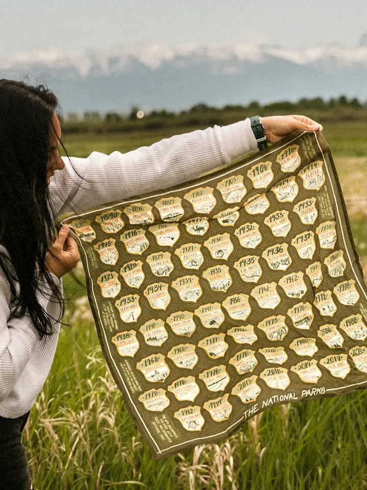 Person holding green beige National Parks bandana showing park badge illustrations and the words The National Parks outside