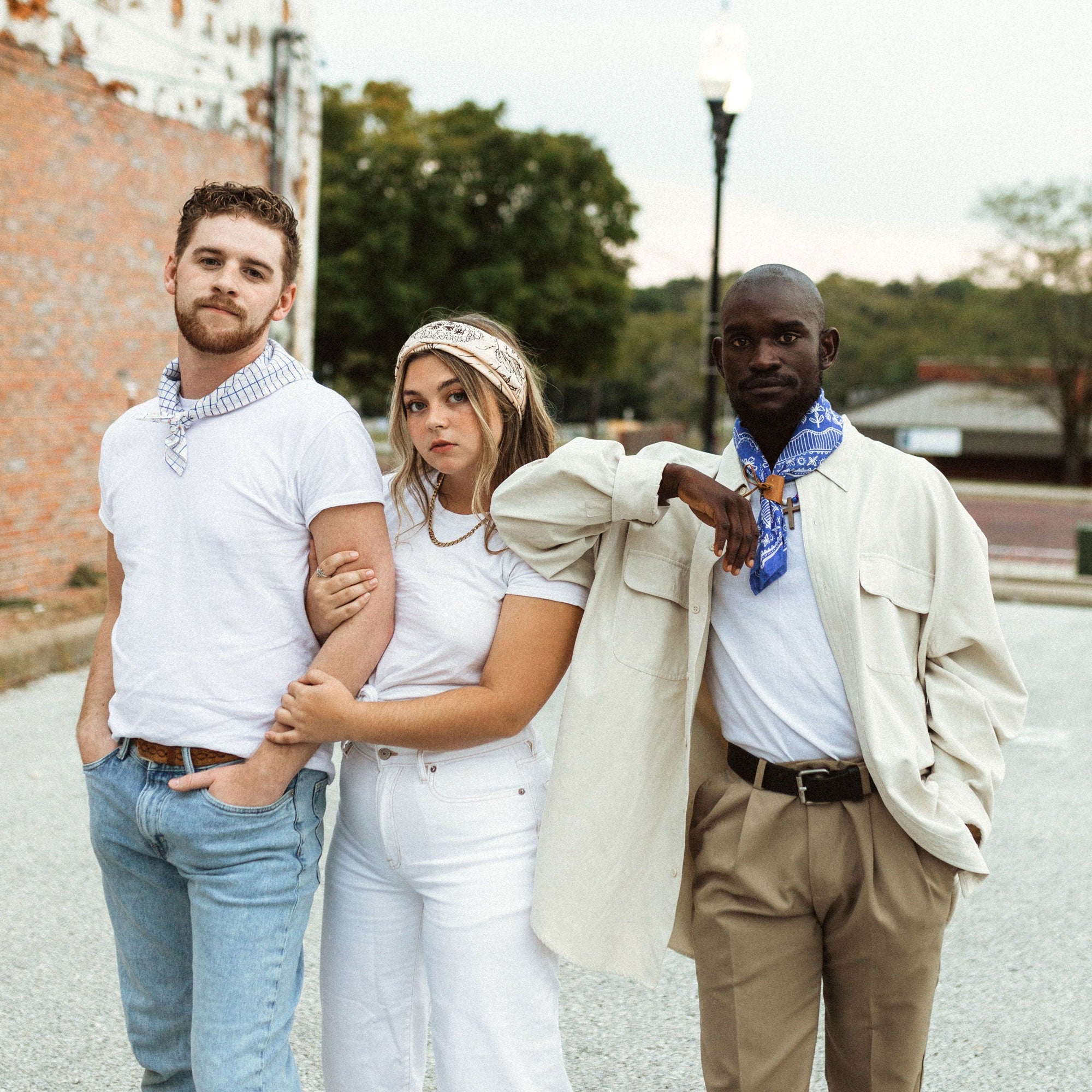 Three people outdoors wearing white tees and bandanas; center with cream snake-print headband, right with blue neck bandana