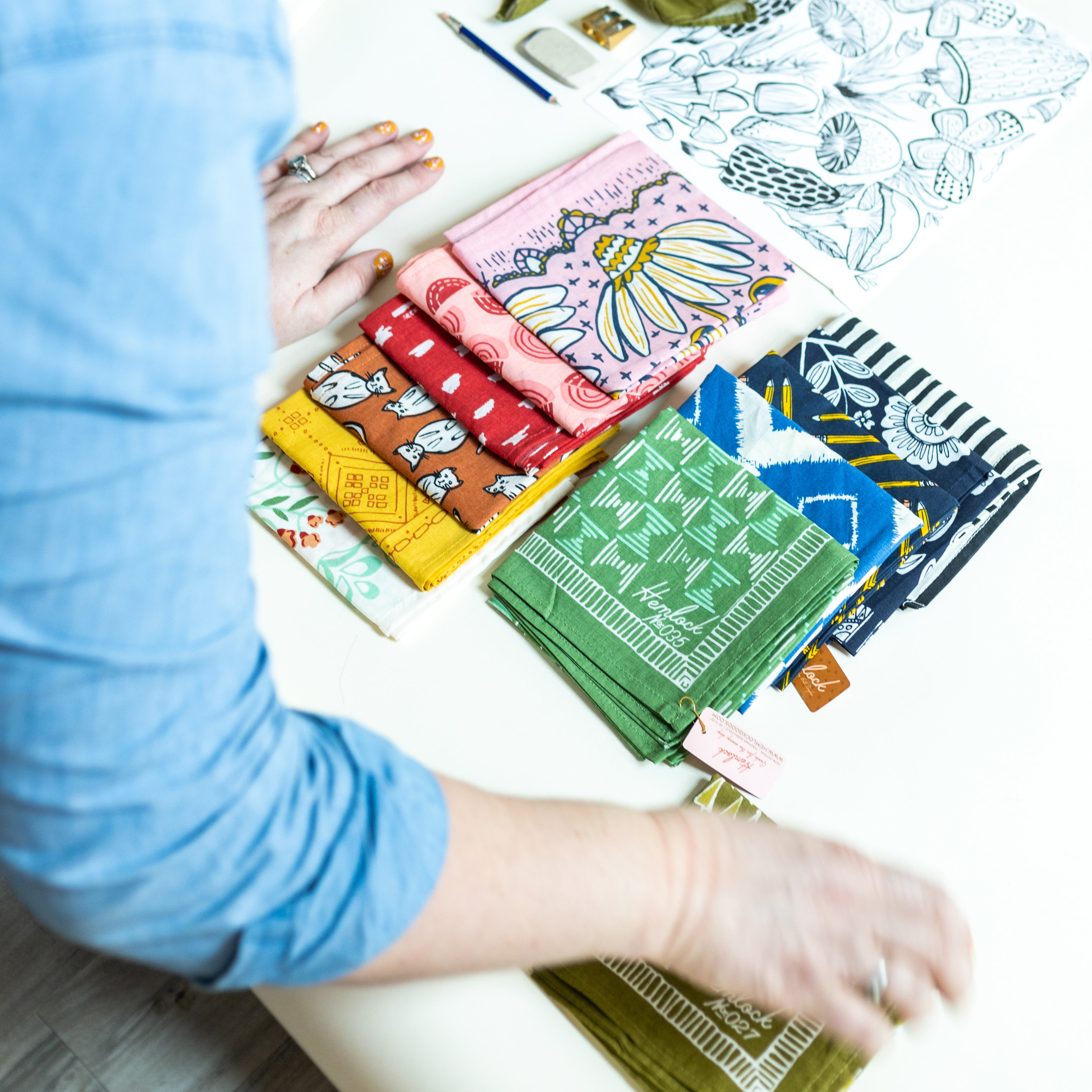 Person arranging stacks of colorful folded bandanas on a white table with sketches and drawing tools nearby