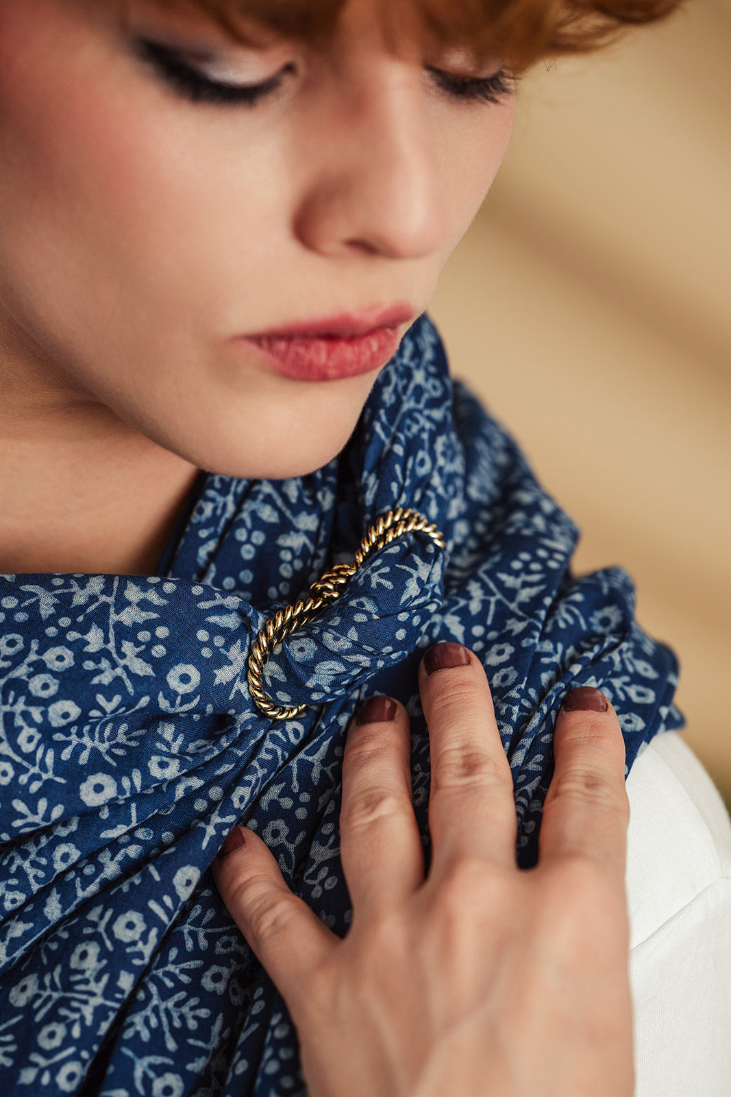 Woman wearing blue patterned scarf fastened with twisted brass rope slide; hand with dark polish touching scarf.