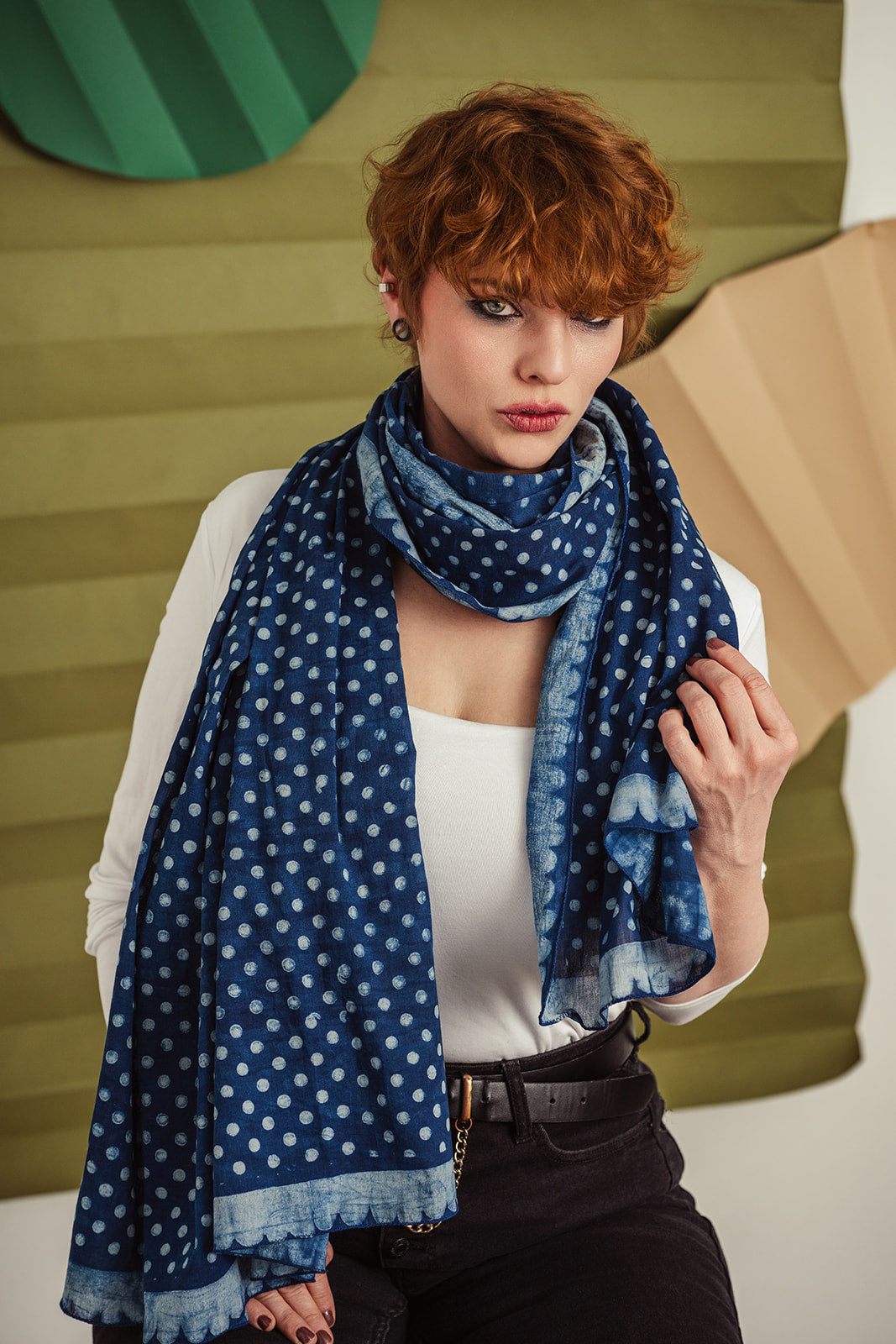 Model wearing navy blue polka-dot scarf draped over a white tank top, seated against layered green-beige backdrop