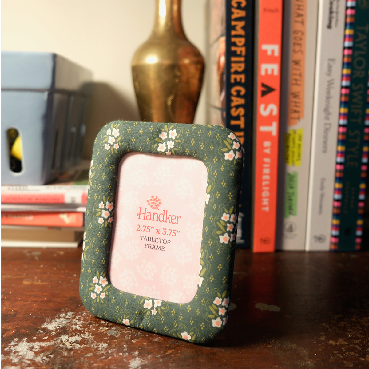 Handker decorative fabric tabletop frame with green floral pattern, standing on a desk with books in the background