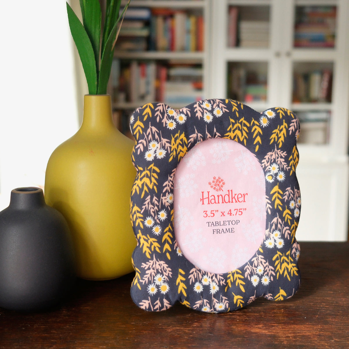 Decorative standing fabric photo frame with navy blue floral design on a wooden surface next to vases and books.