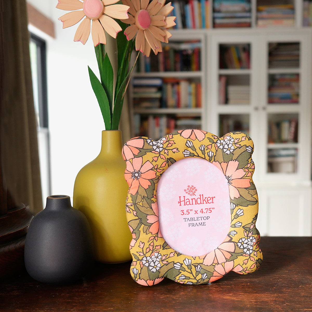 Yellow floral-patterned fabric picture frame with 'Handker' branding on a wooden surface, with a vase and flowers in the background.