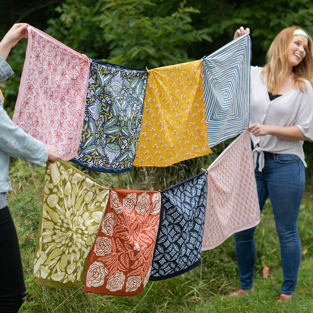 Two people hold a row of colorful patterned bandanas outdoors against a grassy backdrop