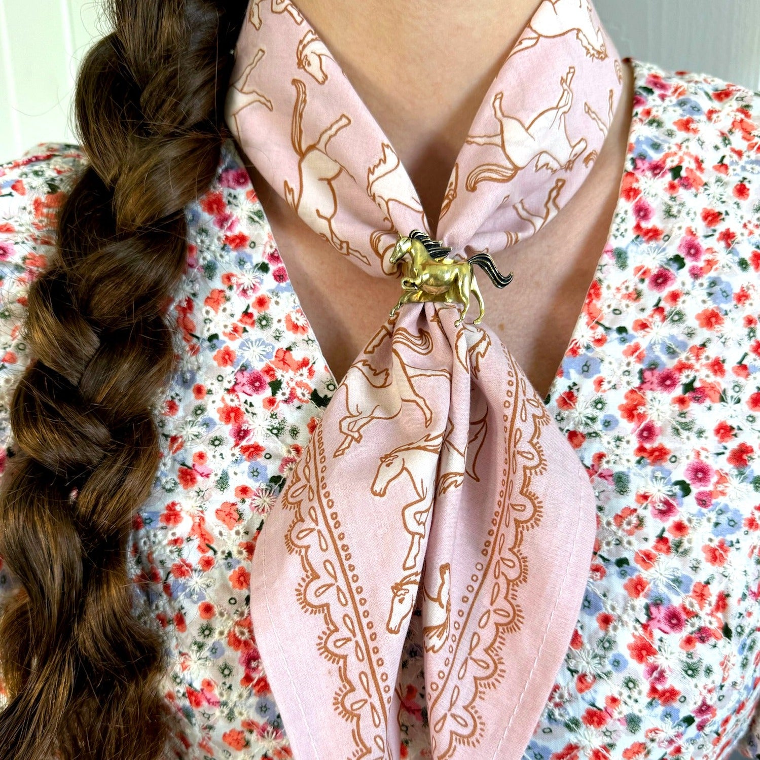 Close-up of a pink patterned scarf with horse design, knotted around neck, and worn over a floral blouse.