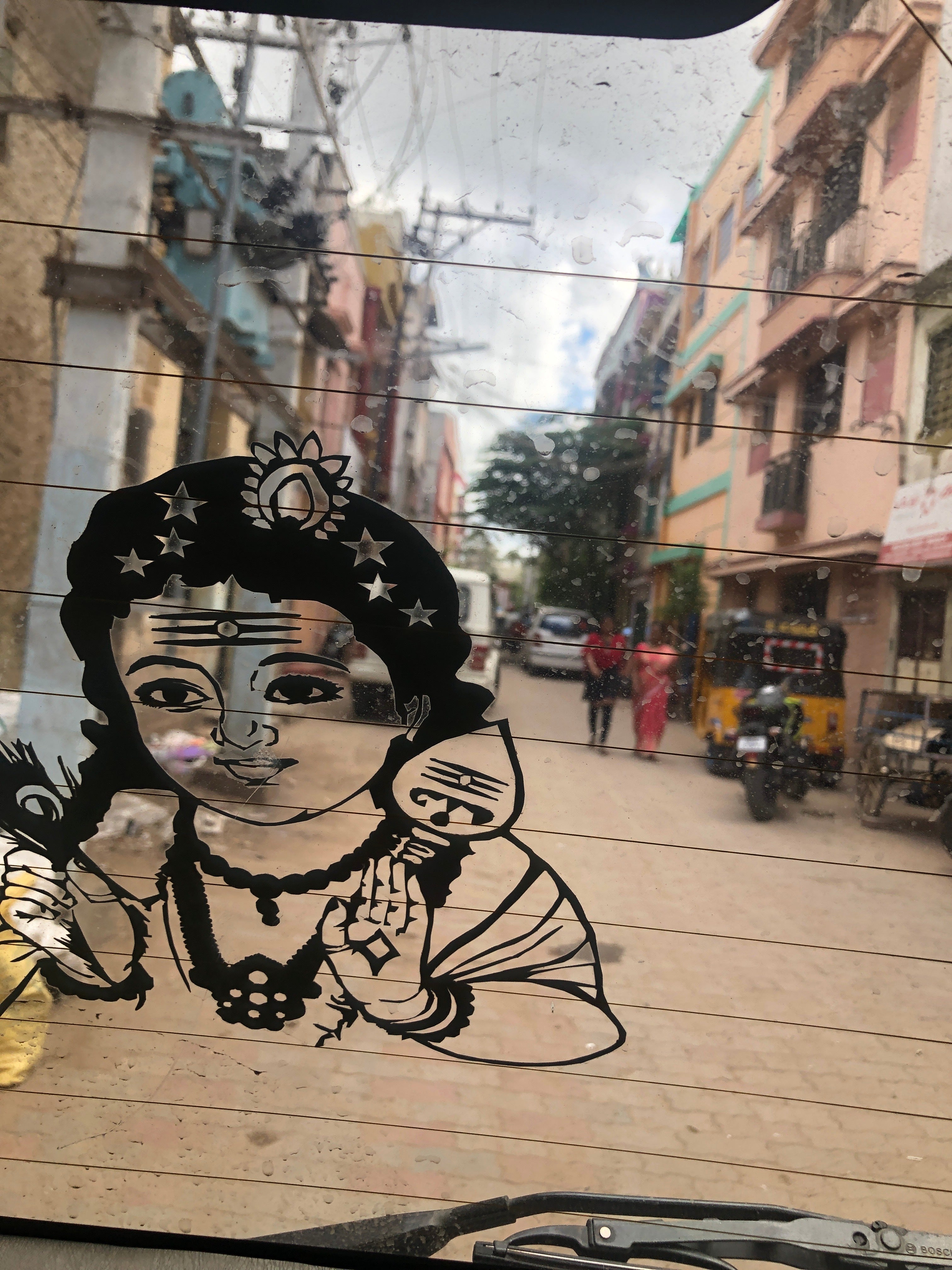 Black decal of baby Krishna on a vehicle rear window looking out to a narrow Indian street with pedestrians and an auto-rickshaw