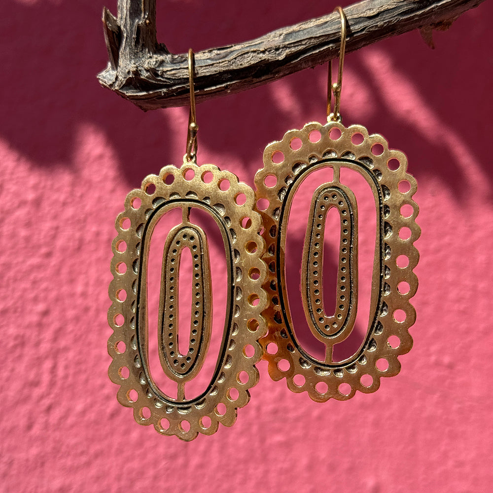 Intricate gold oval earrings hanging on a branch against a pink background.