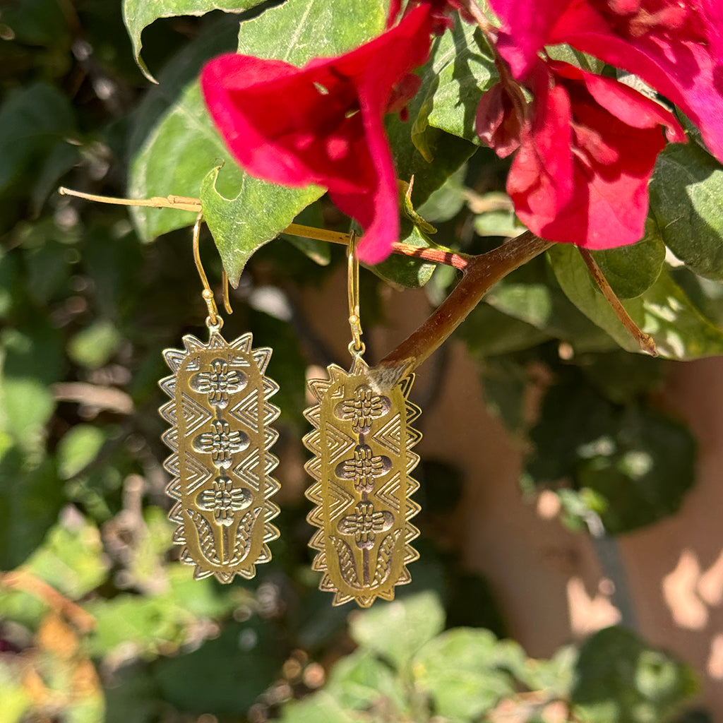 Gold patterned earrings hanging on a plant with red flowers in the background.