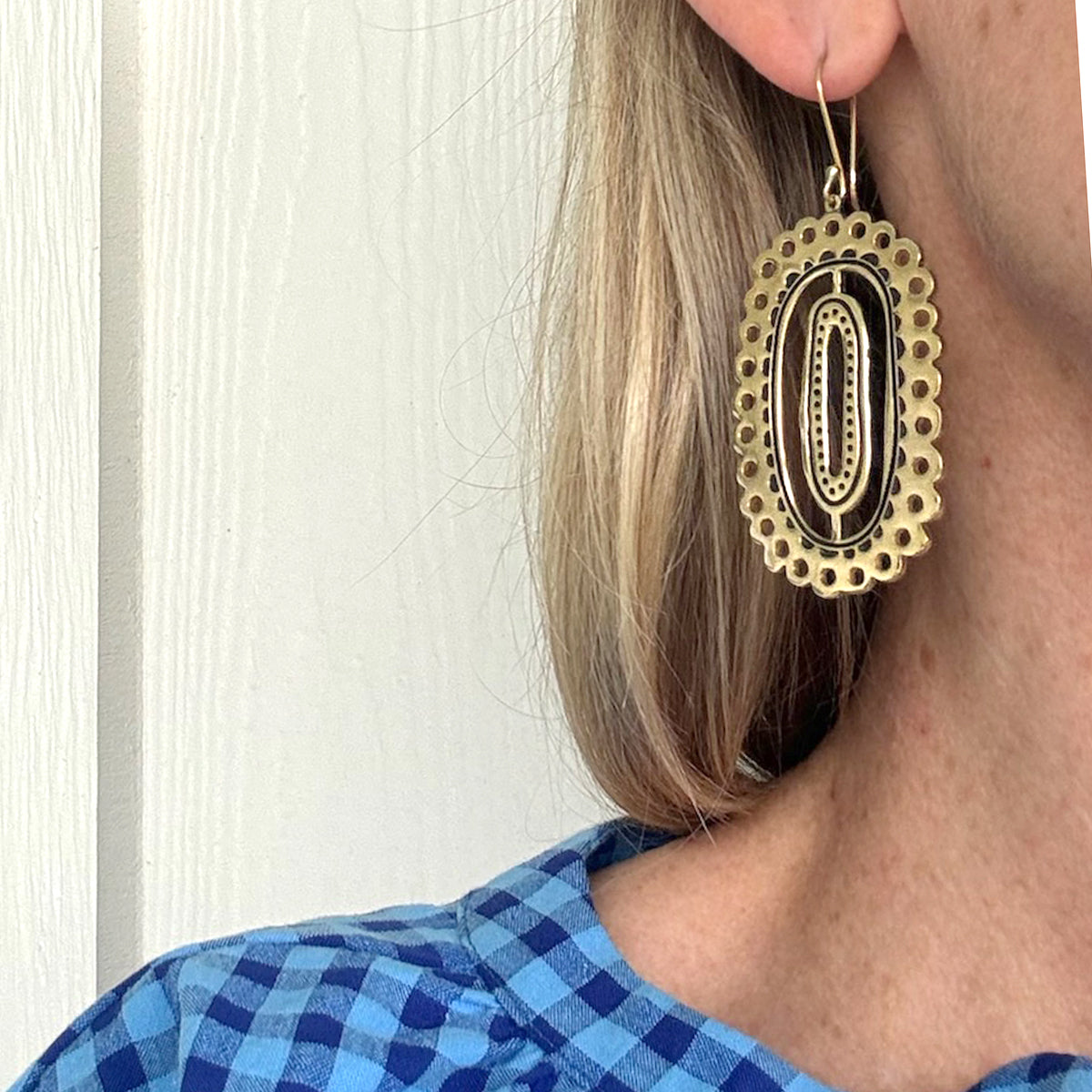 Close-up of a person wearing a large, oval-shaped decorative earring with a blue plaid shirt.