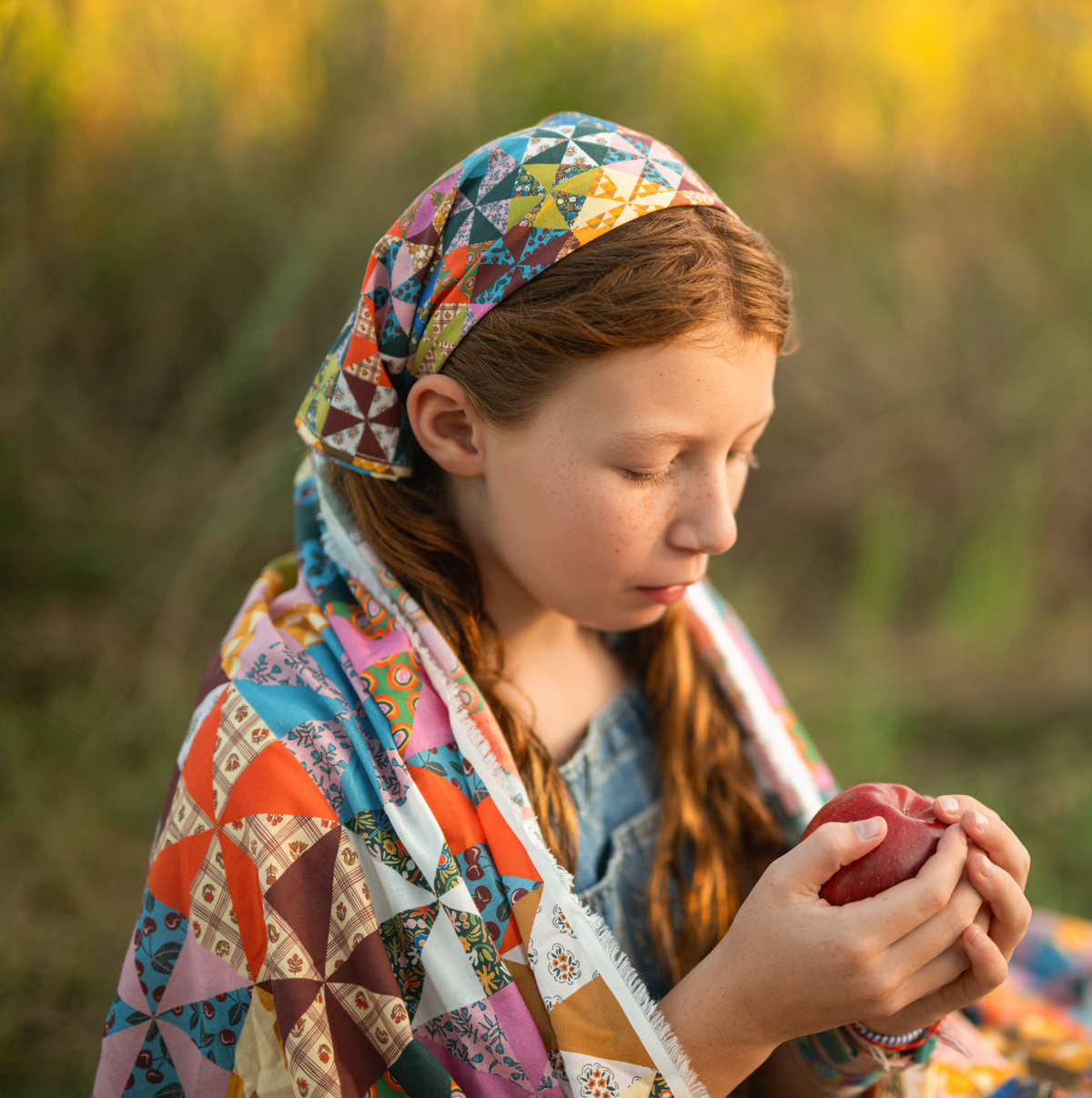 Person wearing colorful Calico fabric as a headscarf and shawl outdoors while holding a red apple