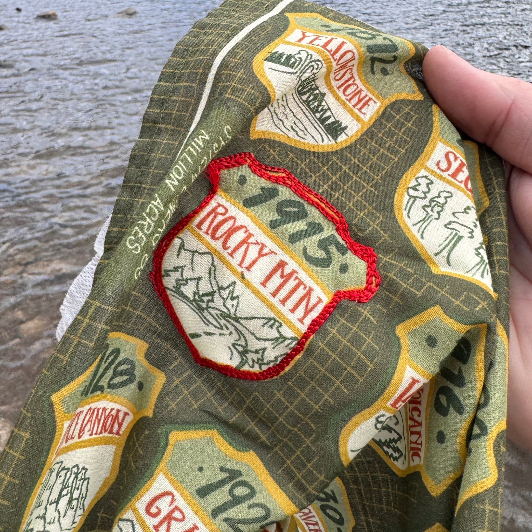 Hand holding green souvenir cloth with park shield patches labeled ROCKY MTN, 1975 and YELLOWSTONE over a river.
