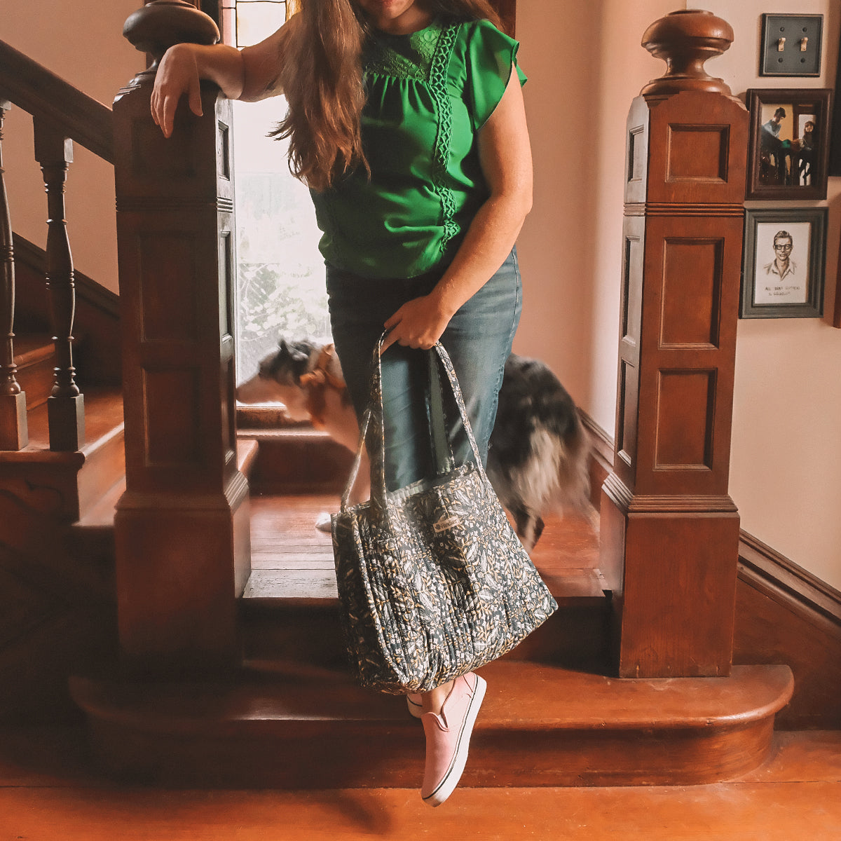 Woman in green top and jeans holding a patterned handbag, standing on wooden stairs with a dog in the background.
