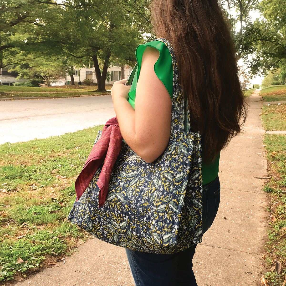 Person with long brown hair carrying a blue floral tote bag over her shoulder, walking on a sidewalk beside grass and trees.