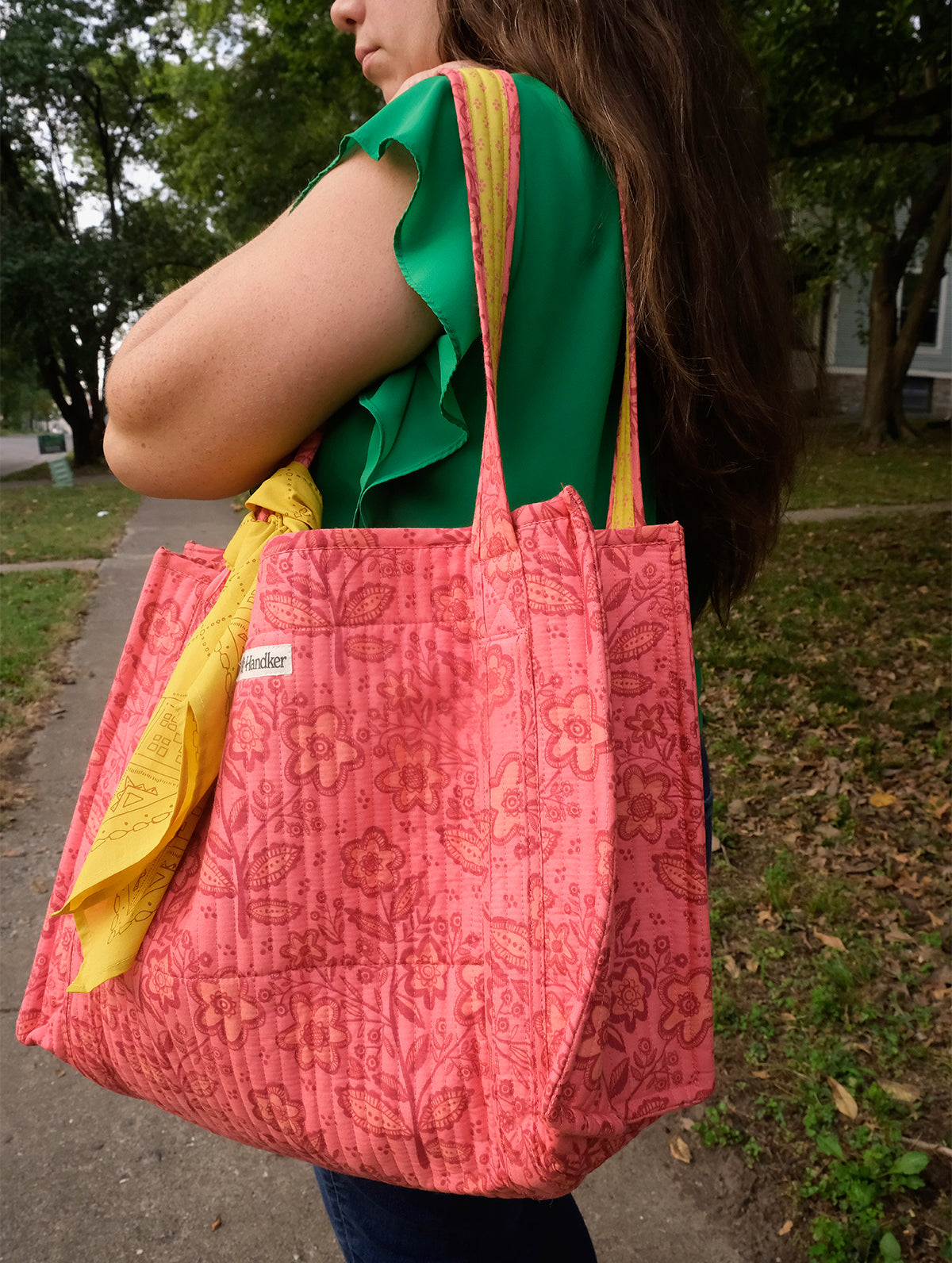 Woman carrying pink patterned tote bag with yellow scarf tied to the handle, walking on a tree-lined sidewalk