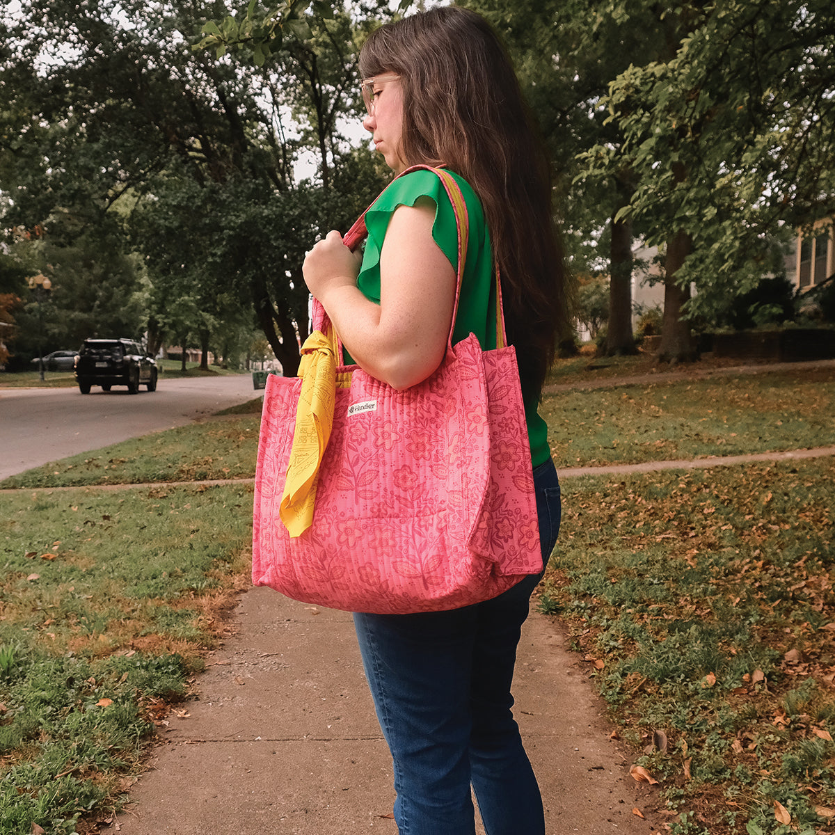 Woman standing on a neighborhood sidewalk holding three reusable shopping bags (pink, yellow, green) over her shoulder