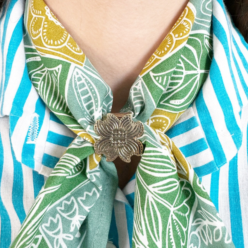 Person wearing a green and yellow floral scarf with a silver flower brooch over a blue and white striped shirt.