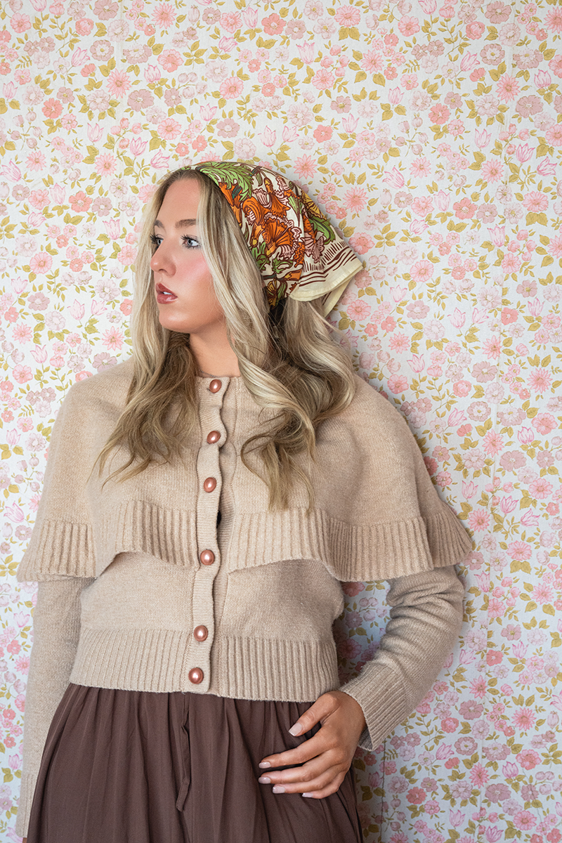 Side view of No. 153 Hannah Bandana tied as headscarf, cream base with orange and green floral print against pink wallpaper