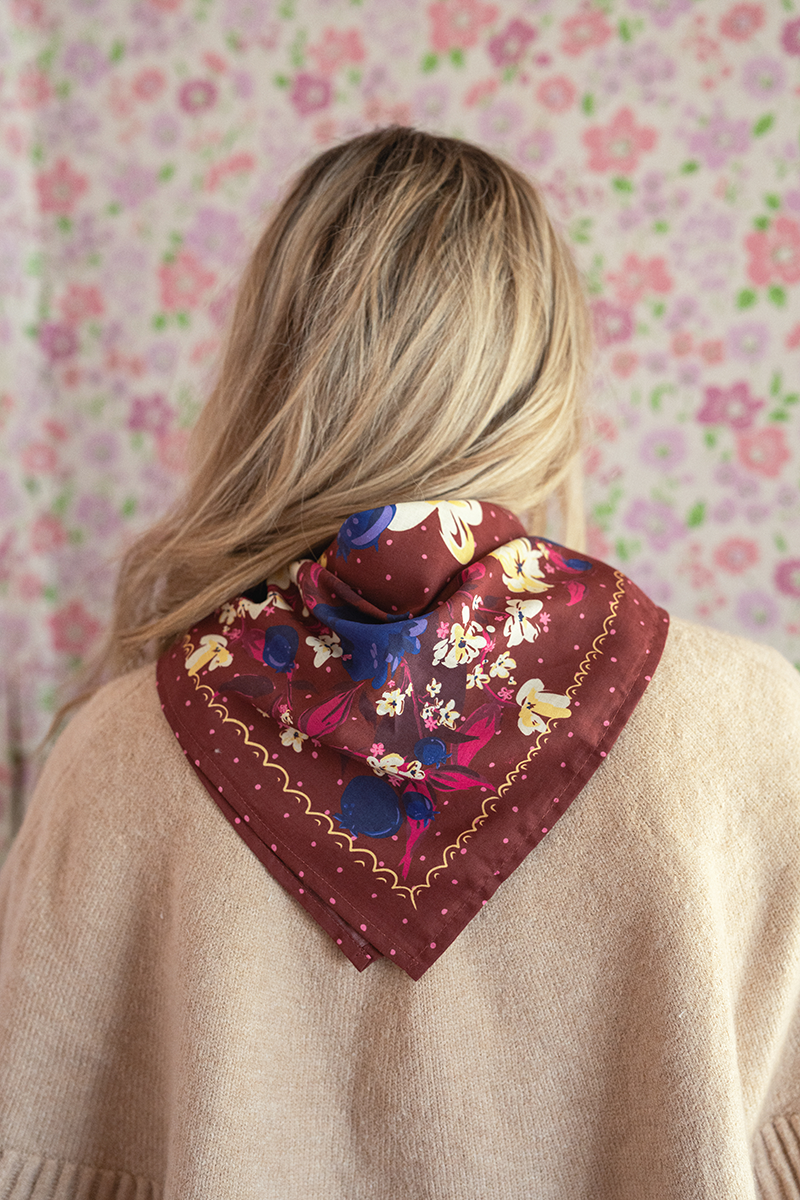 Person wearing a burgundy bandana with a blueberry and floral print around their neck against a floral wallpaper background