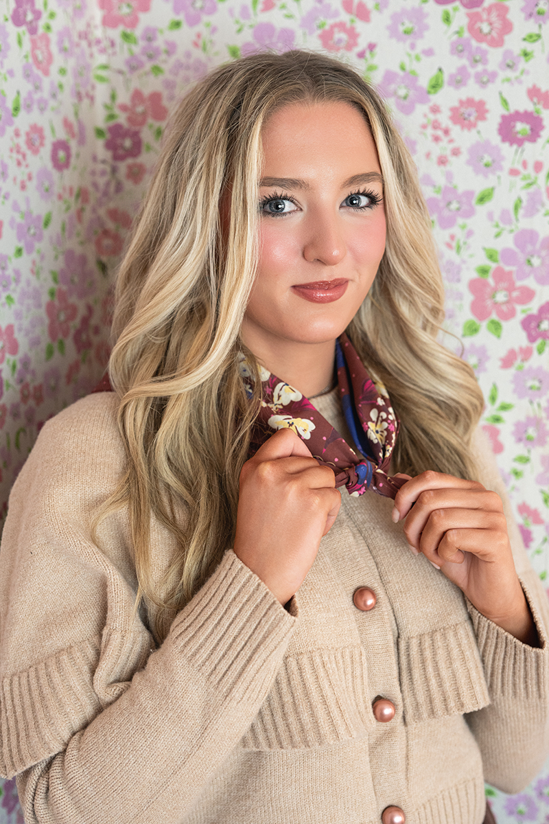 Woman wearing a beige cardigan and burgundy floral patterned bandana against a floral background