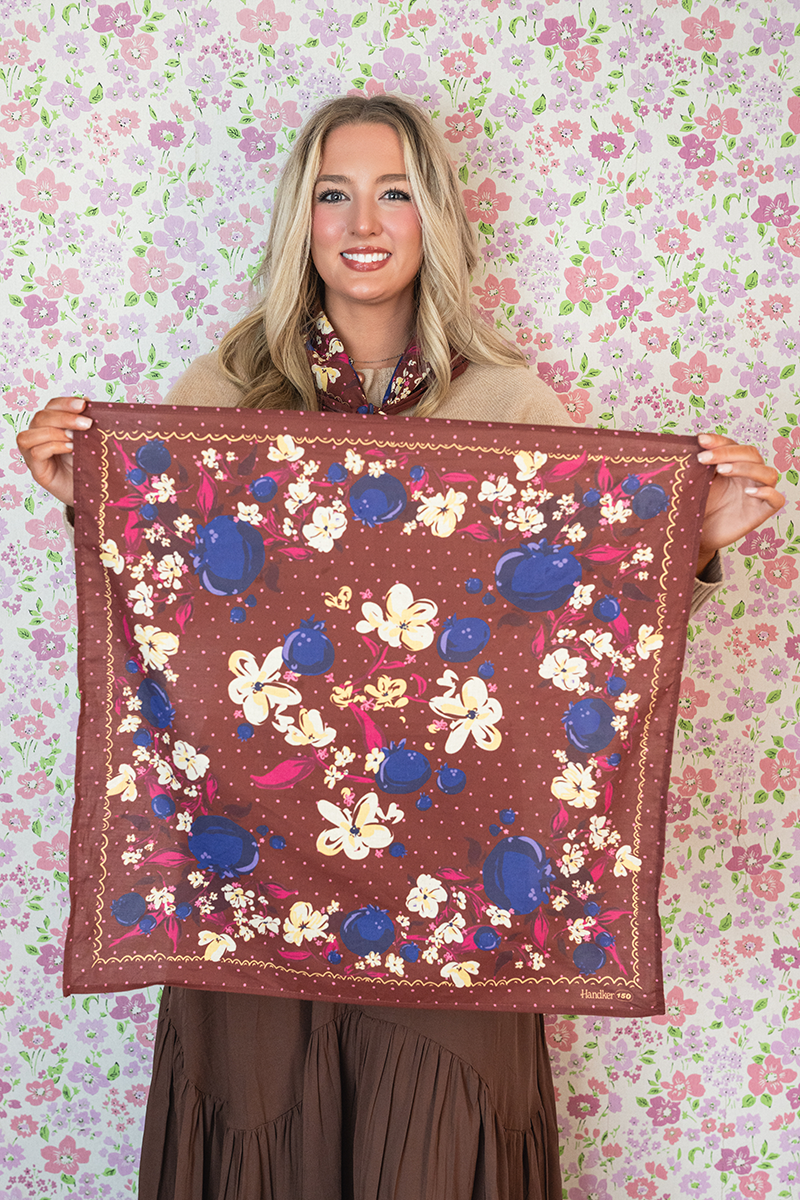Woman holding a burgundy bandana with a polka-dot, blueberry, and floral pattern against a floral background