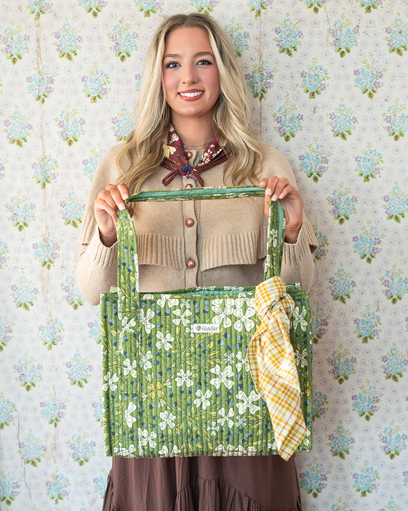 Front view of quilted tote bag with green floral print and a tied yellow plaid bandana held by a model