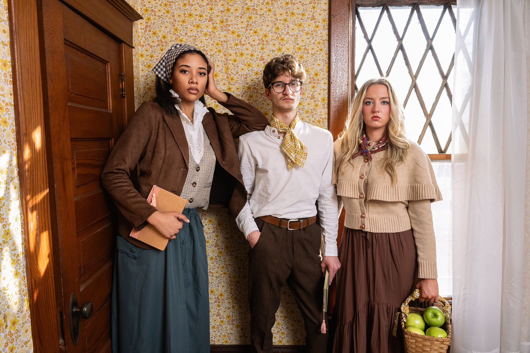Three people in vintage clothing and colorful patterned bandanas standing in a room with patterned wallpaper and a window.