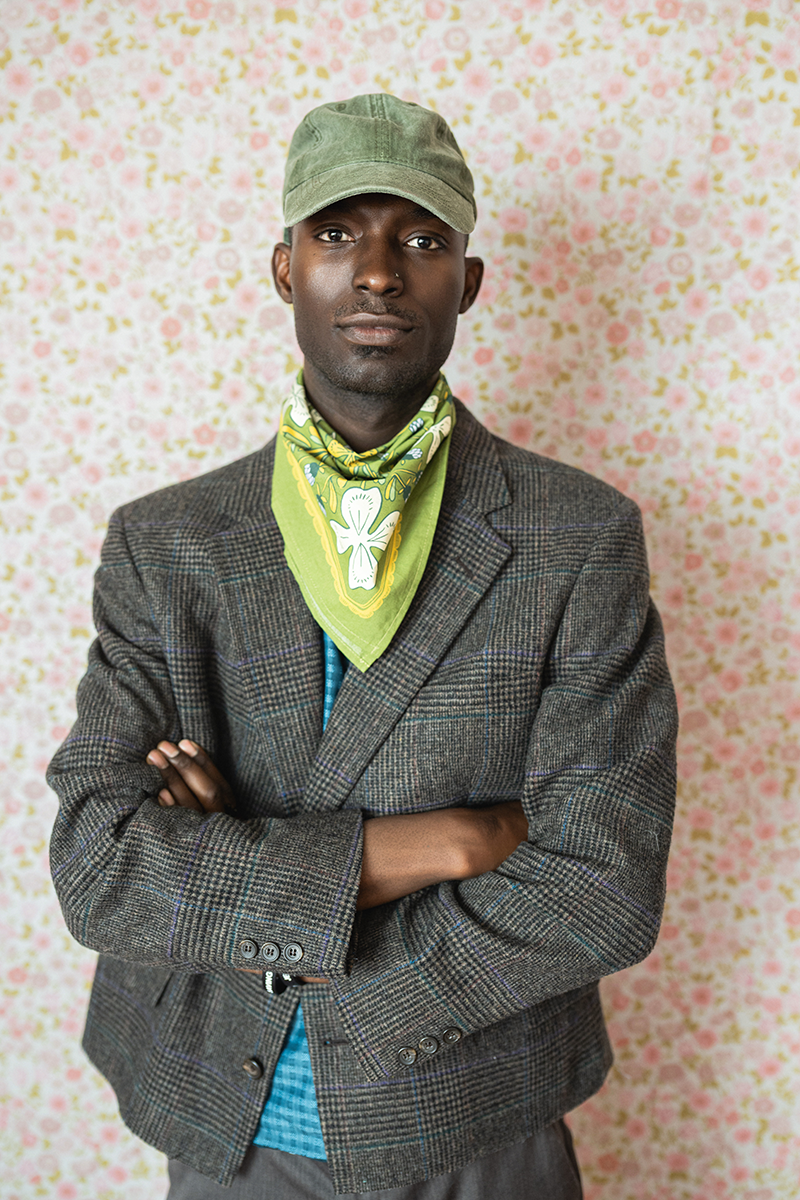 Person wearing a gray blazer, green cap, and green floral bandana against a floral-patterned background