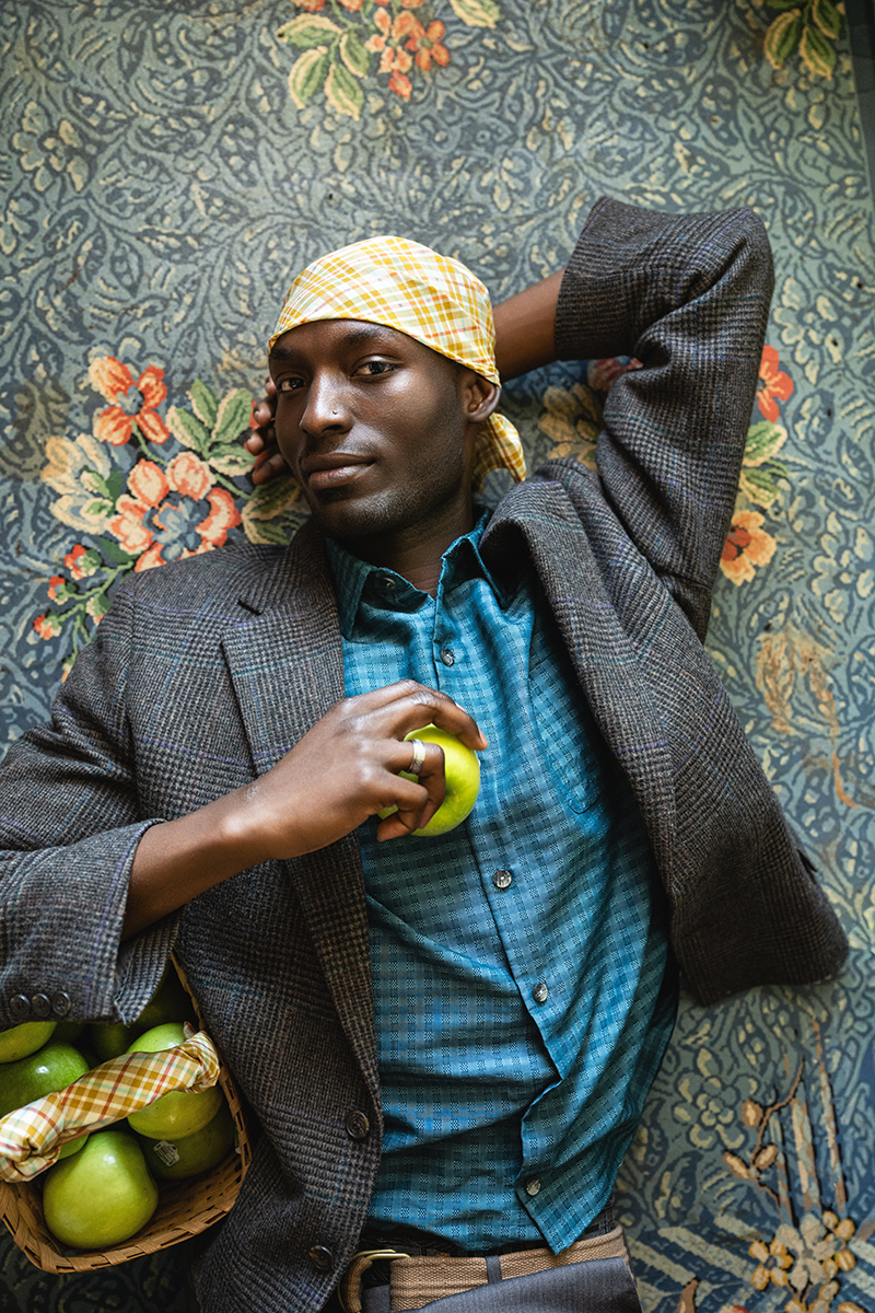 Man with a yellow plaid bandana tied on his head holding a basket of apples against a patterned background