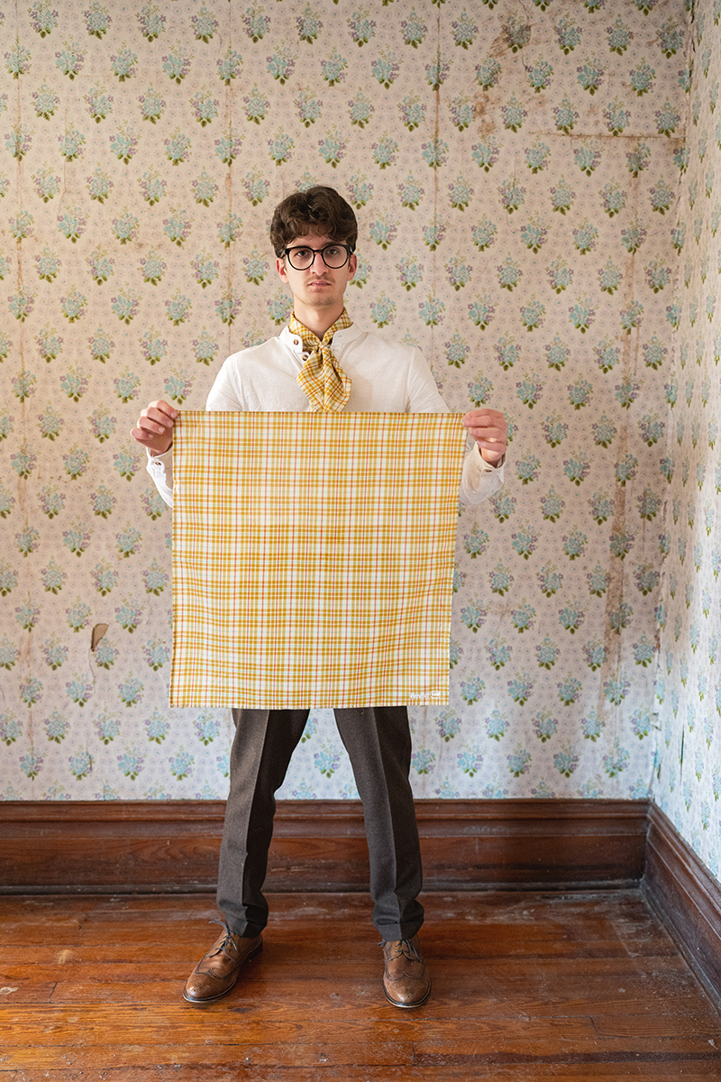 Man holding a yellow checkered bandana against a patterned wall.